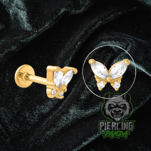 May include: A gold-colored butterfly-shaped earring with clear, sparkling stones. The earring is shown in two views, one with the post and another within a circle. The background is a dark green velvet.