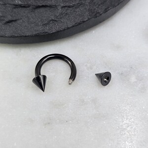 May include: A black circular barbell body jewelry piece featuring a smooth, curved shaft with one attached cone-shaped spike and one detached matching cone-shaped spike. The metal has a glossy finish, and the threaded end of the barbell is visible.