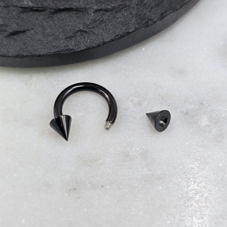 May include: A black circular barbell body jewelry piece featuring a smooth, curved shaft with one attached cone-shaped spike and one detached matching cone-shaped spike. The metal has a glossy finish, and the threaded end of the barbell is visible.