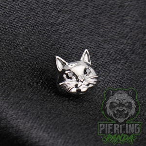 May include: A silver-toned cat head stud earring featuring two small black stones set as eyes. The cat design includes detailed engraved whiskers and pointed ears, presented with a polished metallic finish on a dark, textured fabric surface.
