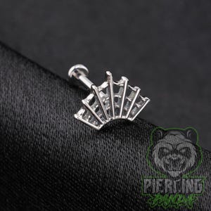 May include: A silver-toned labret stud featuring a decorative top shaped like a fan with five distinct vertical segments. The jewelry piece has a smooth, metallic finish and a straight post extending from the back of the fan-shaped design.
