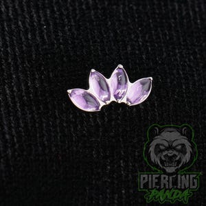 May include: A silver-toned body jewelry piece featuring four marquise-cut purple amethyst stones arranged in a fan-like crown shape. The stones are set in a metallic frame, creating a curved, symmetrical design suitable for various piercings.