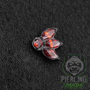 May include: A black metal body jewelry piece featuring a cluster of four faceted red gemstones. The design includes one small round stone at the top and three marquise-shaped stones arranged in a fan-like pattern, all set in a dark metal frame.
