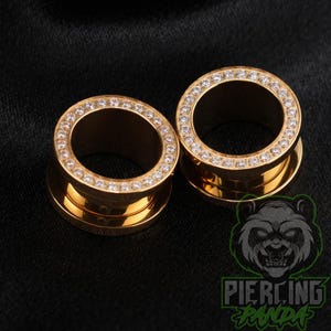 May include: A pair of gold-toned ear tunnels featuring a circular front face encrusted with a single row of clear, sparkling round-cut stones. The tunnels have a smooth, polished metallic finish and a hollow centre, designed for stretched ear piercings.