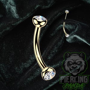 May include: A gold-colored curved barbell body piercing with a clear, round gemstone at each end. The jewelry is displayed against a dark green velvet background. A smaller, detached barbell is also visible.