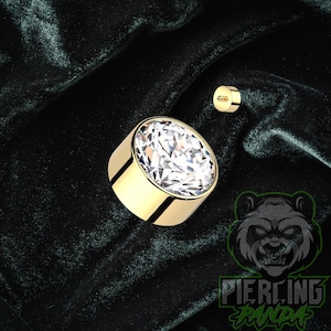 Peut inclure: Un bijou de piercing corporel rond de couleur or avec une grosse gemme claire et facettée. Un petit bijou assorti est également visible. Les bijoux sont posés sur un fond vert foncé. Le logo Piercing Panda est en bas à droite.