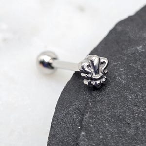 May include: Silver barbell with a fleur-de-lis design on one end and a ball on the other end.