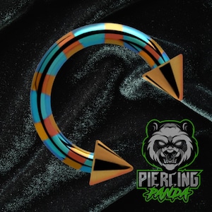 May include: A colorful circular barbell piercing with orange, blue, and black stripes. The ends are gold with black triangles. The logo "Piercing Panda" is in the bottom right corner.
