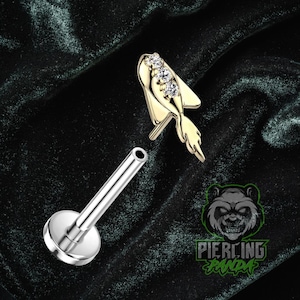 Puede incluir: Un piercing labial dorado con tres gemas transparentes y un diseño en forma de pez. El piercing se muestra con un poste plateado separado y una base plana y redonda. La imagen también incluye un logotipo con un oso y las palabras "Piercing Panda."