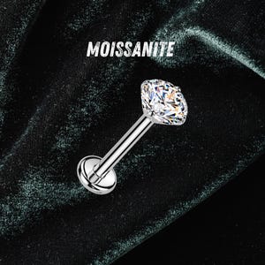 May include: A silver-toned labret stud body jewelry piece featuring a round, faceted, clear moissanite gemstone set in a prong setting at one end and a flat circular disc base at the other. The word MOISSANITE is printed above the jewelry.