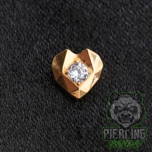 May include: A gold-toned heart-shaped jewelry piece featuring a faceted geometric surface and a single clear round gemstone set in the center. The design has sharp, angular edges and a polished metallic finish, creating a distinct multi-faceted look.