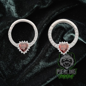 May include: Two silver-colored hinged hoop earrings. Each earring features a heart-shaped, red gemstone pendant with a surrounding halo of small, clear gemstones. The hoops are also lined with clear gemstones. The background is dark green.
