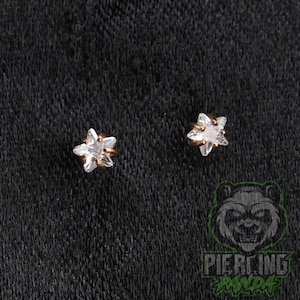 May include: A pair of stud earrings featuring a five-pointed star design. Each earring is crafted from a gold-toned metal and is set with a clear, faceted gemstone in the center, creating a bright, reflective appearance for these small accessories.