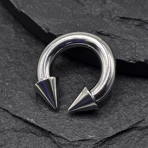 Pode incluir: Uma barra circular prateada com duas pontas pontiagudas. A barra é feita de aço cirúrgico e é projetada para piercings corporais.