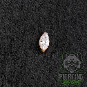May include: A marquise-cut clear gemstone set in a gold-toned metal prong setting. The piece features a pointed oval shape with multiple facets that reflect light, creating a bright appearance. The jewelry component is designed for body piercing.