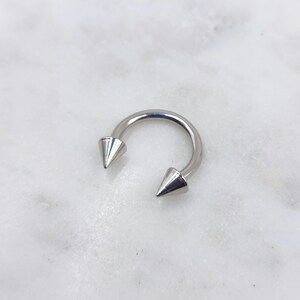May include: A silver horseshoe-shaped piercing with conical spikes at each end. The jewellery is set against a white marble background, highlighting the metallic sheen of the piercing.
