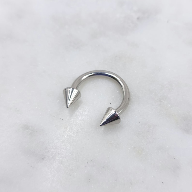 May include: A silver horseshoe-shaped piercing with conical spikes at each end. The jewellery is set against a white marble background, highlighting the metallic sheen of the piercing.