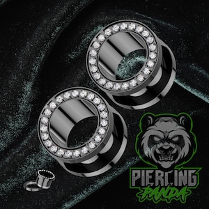 May include: Two black tunnel ear gauges with a ring of clear crystals around the outer edge. A smaller gauge and the Piercing Panda logo are also visible. The gauges are on a dark green background.
