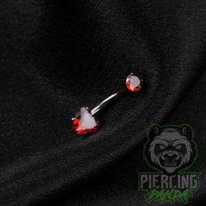 May include: A curved barbell belly button ring featuring a large heart-shaped red gemstone at one end and a smaller round red gemstone at the other end, both set in a polished silver-toned metal base.