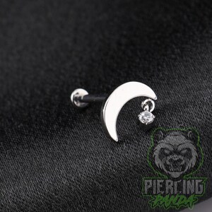 May include: A silver-toned metal body jewelry stud featuring a crescent moon shape. A small, clear, round faceted gemstone is suspended from the lower tip of the moon. The stud includes a straight post with a flat circular backing for secure placement.