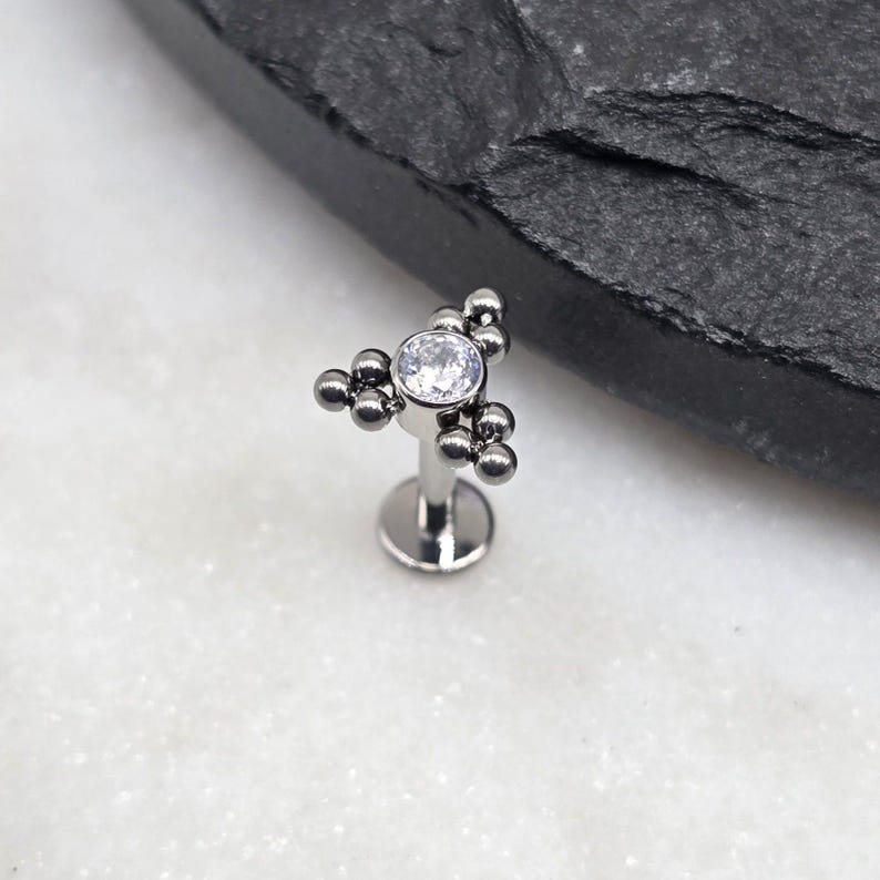 May include: A silver body piercing with a clear gem and a cluster of small silver beads in a triangular shape.