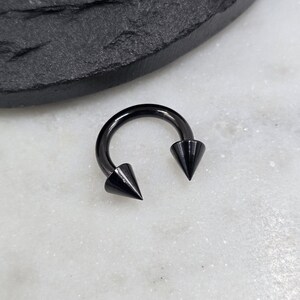 May include: A circular horseshoe-shaped body jewelry piece with a smooth, polished black finish. The ends of the curved bar feature two sharp, conical spikes that point inward toward each other, creating a distinct and symmetrical appearance.