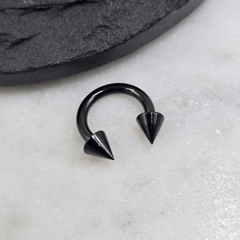 May include: A circular horseshoe-shaped body jewelry piece with a smooth, polished black finish. The ends of the curved bar feature two sharp, conical spikes that point inward toward each other, creating a distinct and symmetrical appearance.
