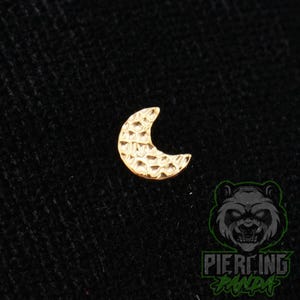 May include: A small, gold-colored crescent moon stud with a textured, hammered surface finish. The piece features a distinct cratered pattern across its curved face, creating a reflective appearance. The item is presented against a dark background.