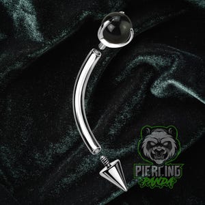 May include: A silver-colored curved barbell body piercing jewelry with a black ball on one end and a silver cone on the other. The jewelry is disassembled, showing the threaded ends. The background is a dark green velvet.