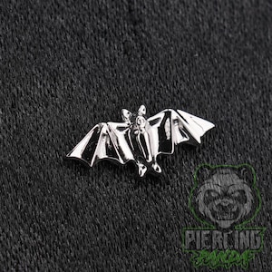 May include: A silver-toned metal bat-shaped jewelry piece featuring a polished finish with defined wings and a central body. The bat is depicted with its wings spread wide in a horizontal orientation, showcasing smooth surfaces and sharp edges.