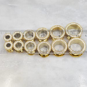 May include: A collection of gold-colored ear gauges with clear, sparkling rhinestones around the outer edge. The gauges are arranged in a row, with a ruler placed next to them for size comparison. The gauges vary in size.