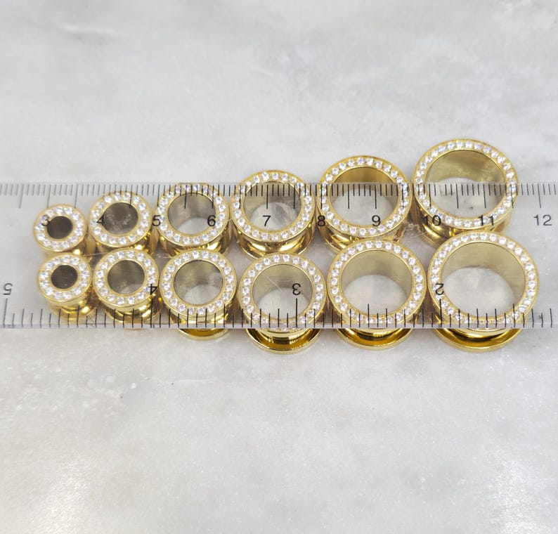 May include: A collection of gold-colored ear gauges with clear, sparkling rhinestones around the outer edge. The gauges are arranged in a row, with a ruler placed next to them for size comparison. The gauges vary in size.