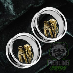 May include: A pair of silver-colored ear gauges with a detailed gold-colored elephant design inside. The elephant has intricate detailing on its body and trunk. The gauges are set against a dark green background.