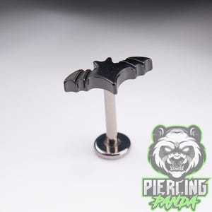 May include: A dark metallic labret stud featuring a flat bat-shaped top with scalloped wing edges. The jewelry consists of a straight post and a circular flat back disc, all finished in a polished, dark gunmetal tone with a smooth, reflective surface.