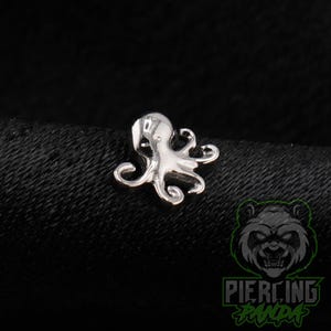 May include: A small, silver-toned metal body jewelry piece shaped like an octopus. The charm features a smooth, reflective surface with detailed tentacles curling outward from the central head, designed for use as a stud or decorative piercing accessory.