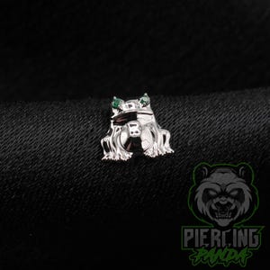 May include: A silver-toned metal body jewelry piece shaped like a frog. The frog features a smooth, polished finish and two small, vibrant green gemstones set as eyes. The design captures the frog in a seated position with visible limbs.