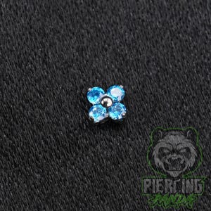 May include: A flower-shaped body jewelry attachment featuring four round, bright blue faceted stones arranged around a central silver-toned metal bead. The piece has a metallic base and is designed for use as a decorative piercing component.