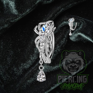 May include: A silver-colored body piercing jewelry set featuring a spider design with clear and blue gemstones. The spider has a dangling teardrop-shaped gem. A smaller matching piece is also shown.
