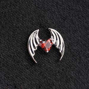 May include: A silver-toned metal charm featuring a central heart-shaped red gemstone flanked by two symmetrical bat-like wings. The piece has a polished metallic finish and a detailed wing structure that curves downward around the red stone.