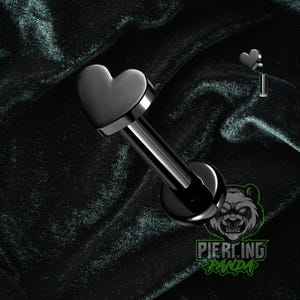 May include: A black heart-shaped labret stud with a flat back and a long, straight post. The jewelry is made of a shiny, dark metal and is designed for lip or ear piercings. The image also shows a small heart charm.