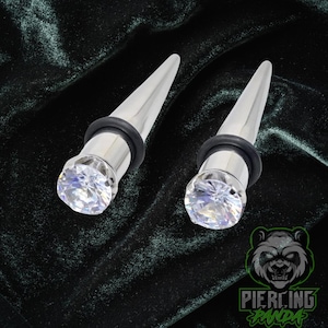 May include: Two silver-colored ear gauges with a clear, faceted gem at the base and a tapered, pointed end. Each gauge has a black rubber ring. The gauges are displayed on a dark green, textured background.