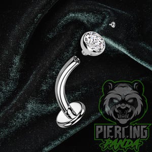 May include: A silver-colored curved barbell body jewelry piece featuring a flat circular base and a detachable top ball set with a clear, faceted round gemstone. The metal surface is polished and reflective, showing a metallic sheen.