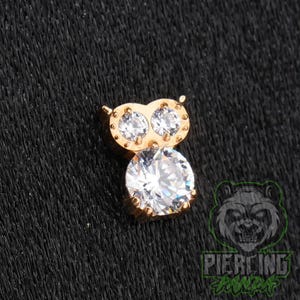 May include: A gold-toned owl-shaped jewelry piece featuring two small clear round stones for eyes and one large clear round stone for the body. The design includes small metal bead accents around the eyes and a textured finish on the metal surface.
