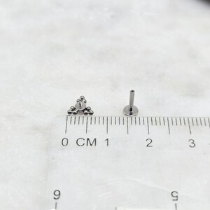 May include: A silver triangle-shaped body jewellery piece with small balls, measured against a ruler in centimetres. The jewellery piece is 1 centimetre wide.