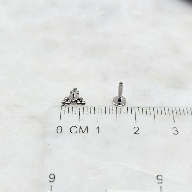 May include: A silver triangle-shaped body jewellery piece with small balls, measured against a ruler in centimetres. The jewellery piece is 1 centimetre wide.