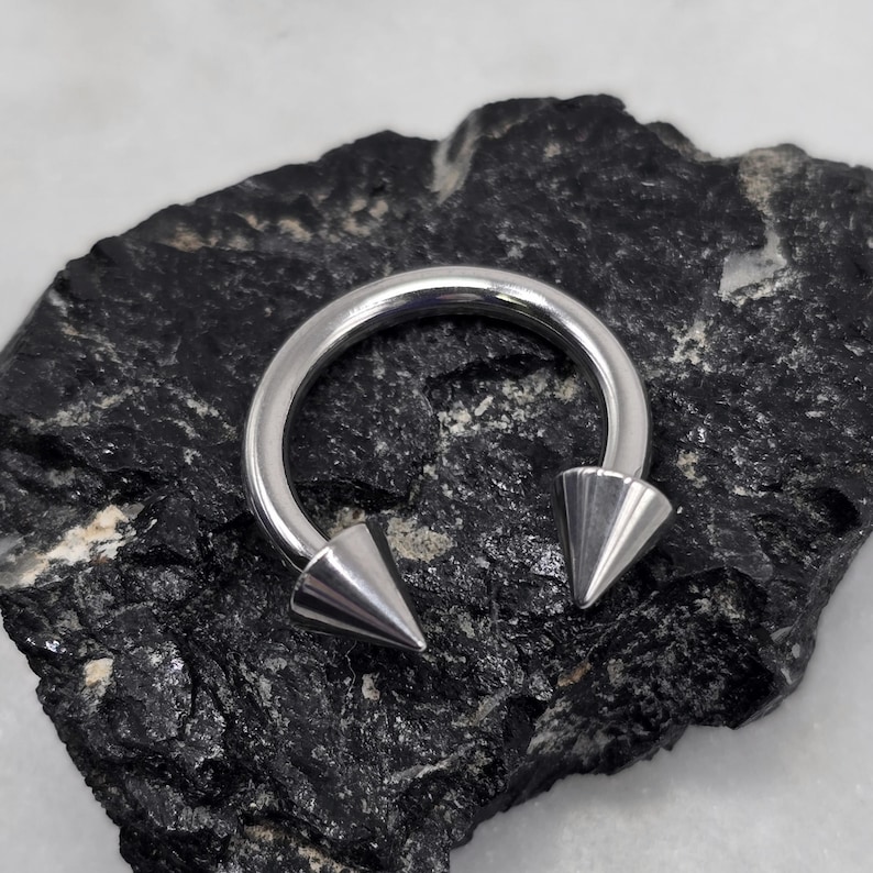 May include: A silver horseshoe piercing with two conical spikes. The jewellery is made of a shiny, metallic material. The piercing is set against a dark, textured background.