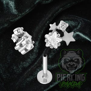 May include: Silver-toned body jewelry with clear cubic zirconia stones. The jewelry includes a star-shaped design, a cluster of stones, and a flat-back post. The background is a dark green fabric.