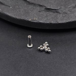May include: A silver body jewellery piercing with a cluster of small balls in a triangular shape.