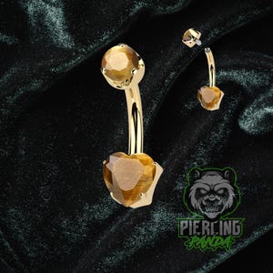Pode incluir: Anel de umbigo dourado com uma pedra preciosa castanha facetada em forma de coração. A barra curva tem uma pedra preciosa a condizer no topo. Uma pequena peça superior a condizer também é mostrada. O fundo é um tecido verde escuro texturizado.