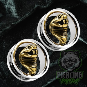 May include: A pair of silver and gold-toned ear gauges featuring a detailed cobra design. The cobra heads are gold, with intricate scale detailing. The gauges have a polished silver outer ring and a gold inner ring.